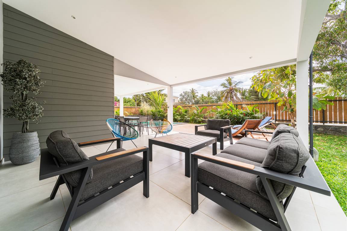 Salon extérieur couvert avec fauteuils et canapé, vue sur la piscine et le jardin