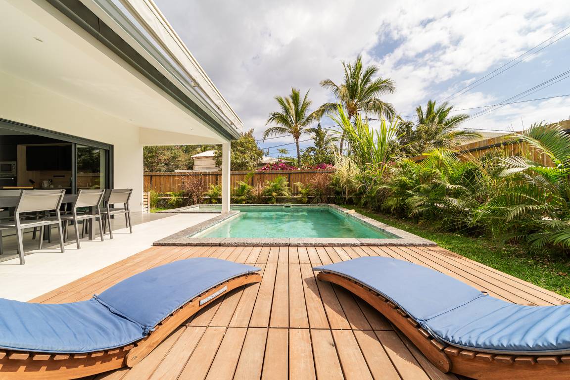 Deux transats aux coussins bleus sur le deck face à la piscine et aux palmiers